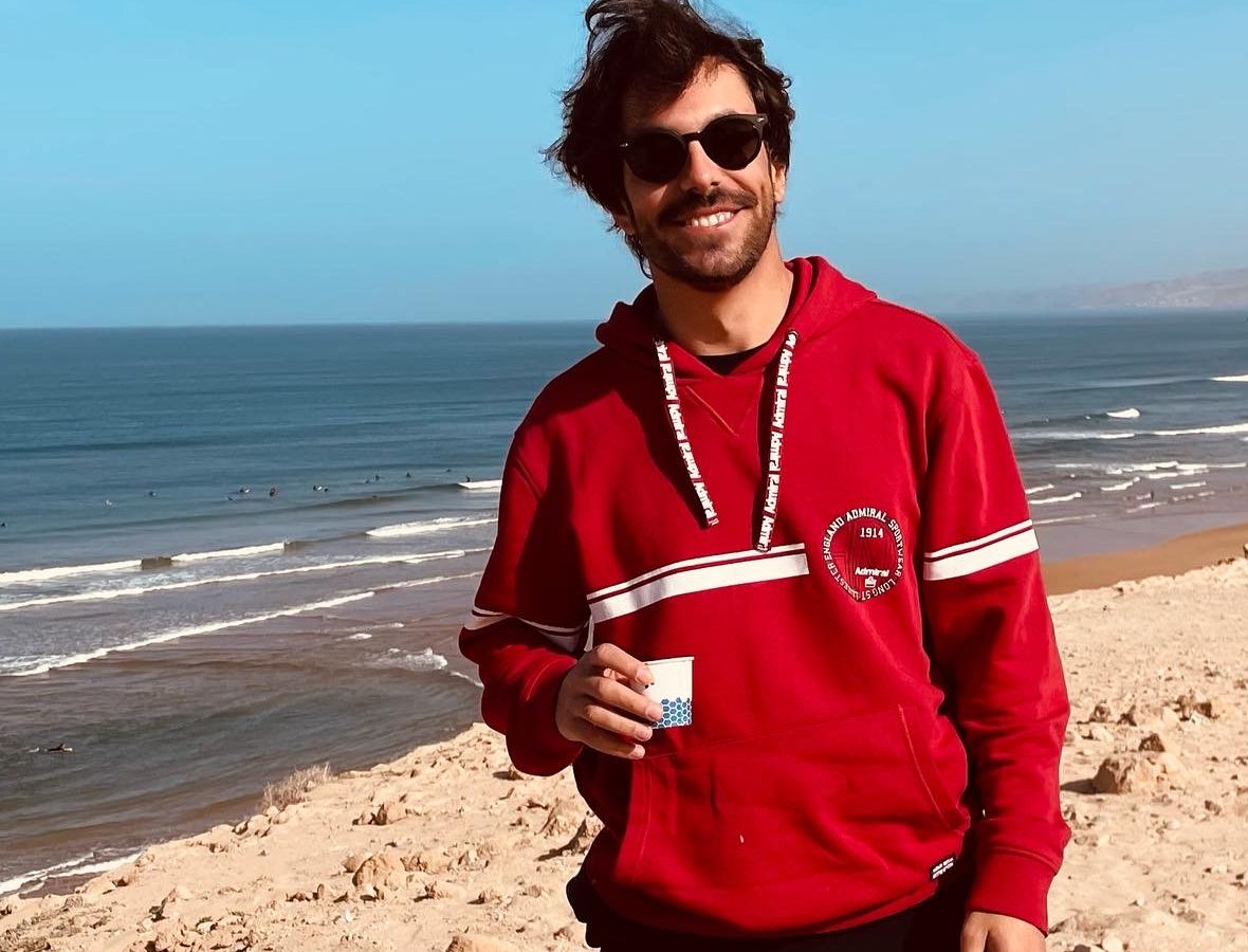 Edoardo su una spiaggia in Marocco, con i surfisti in acqua alle spalle